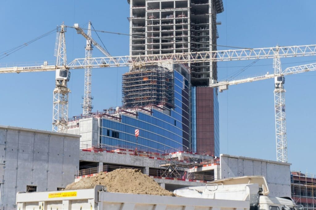 A modern high-rise building with a blue glass facade during its construction phase, with several white tower cranes surrounding it.